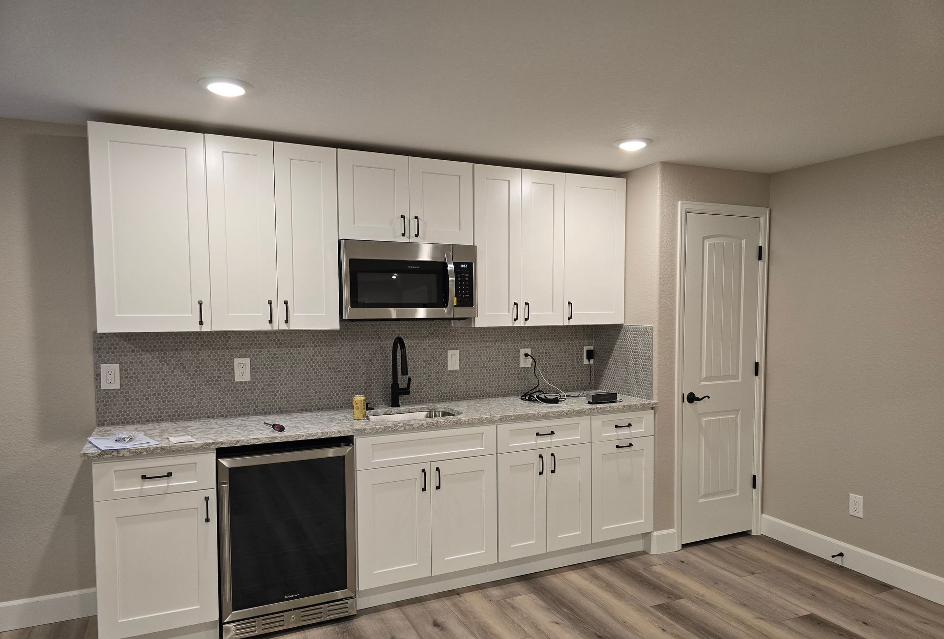 Elegant basement wet bar installation featuring white shaker cabinets, stone countertops, mosaic tile backsplash, undercounter beverage refrigerator, and modern fixtures, enhancing entertainment and hosting capabilities.