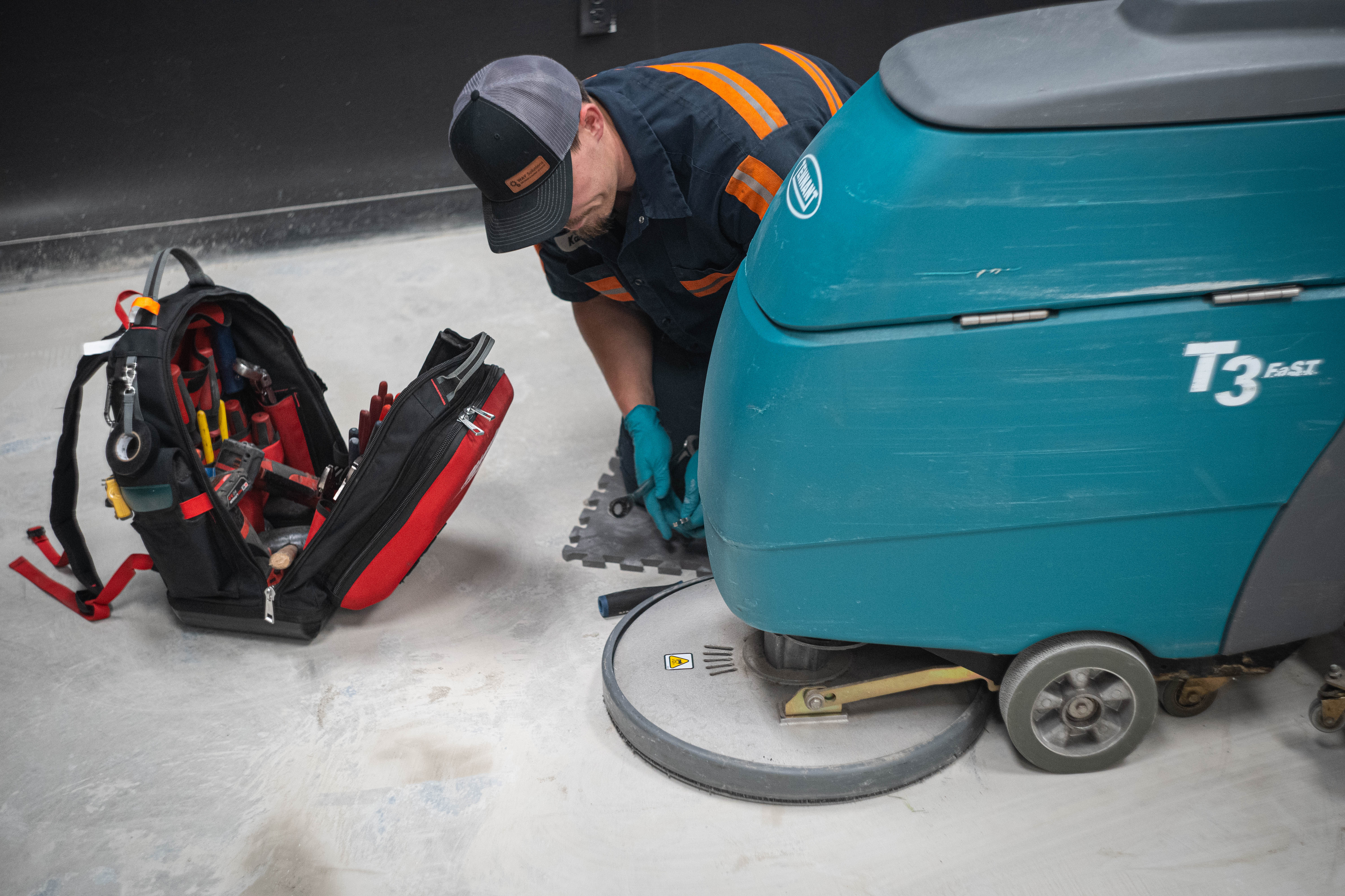 A professional WaySolutions field technician prepares to service the scrub deck of a teal Tennant T3 FAST commercial floor scrubber. With a fully stocked professional tool backpack open and ready, our team ensures that every on-site repair in Layton, Utah, is handled efficiently with the right tools and OEM parts to minimize facility downtime for supermarkets and retail centers.