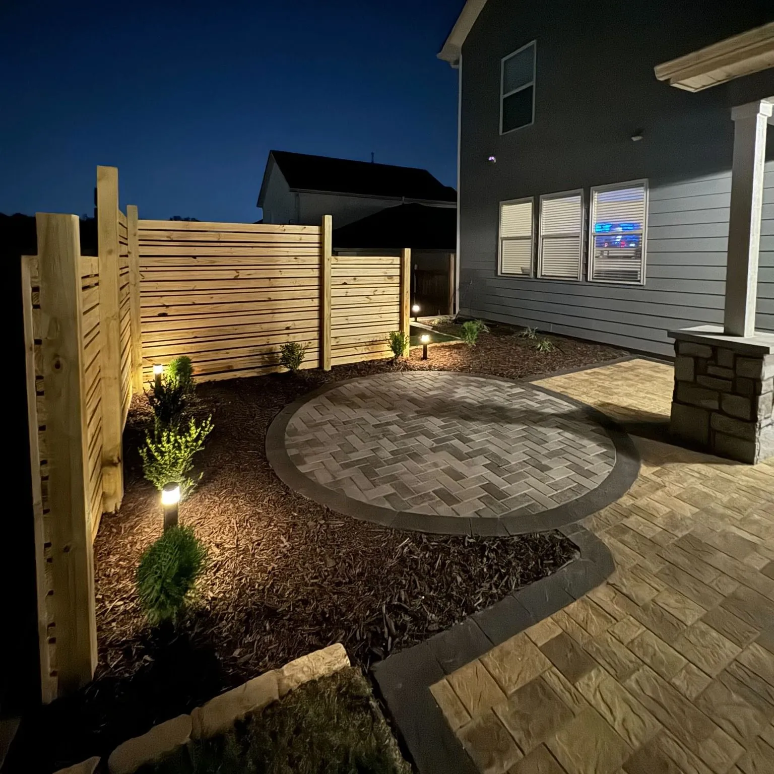 This nighttime view showcases Zamora Design and Build's expertise in creating inviting outdoor spaces with hardscaping, fencing, and strategically placed lights for ambiance and functionality.