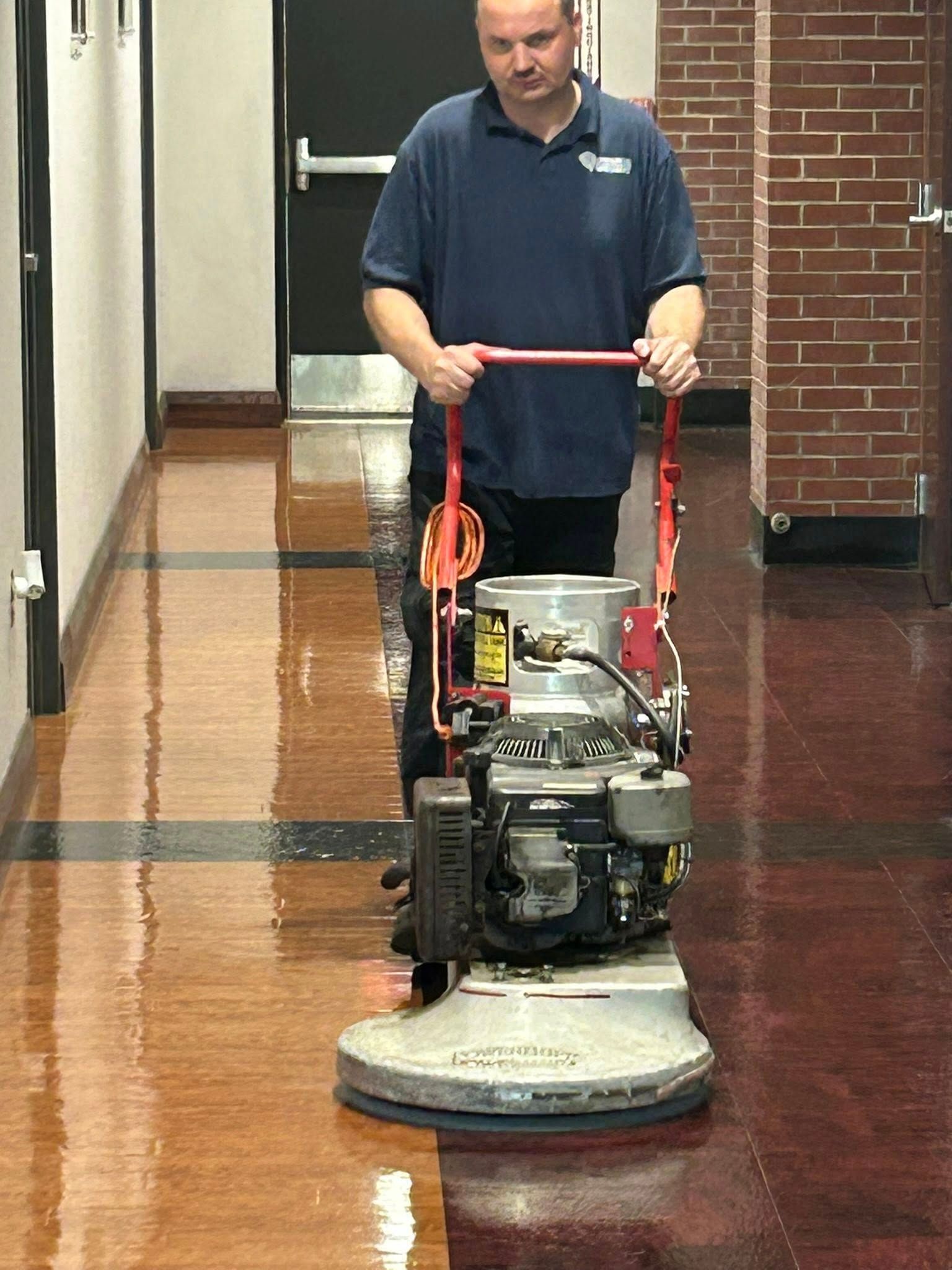 Professional janitor operating industrial floor buffer machine in hallway, polishing surface to restore shine, with visible reflection, brick wall background, and efficient cleaning process in commercial indoor environment.