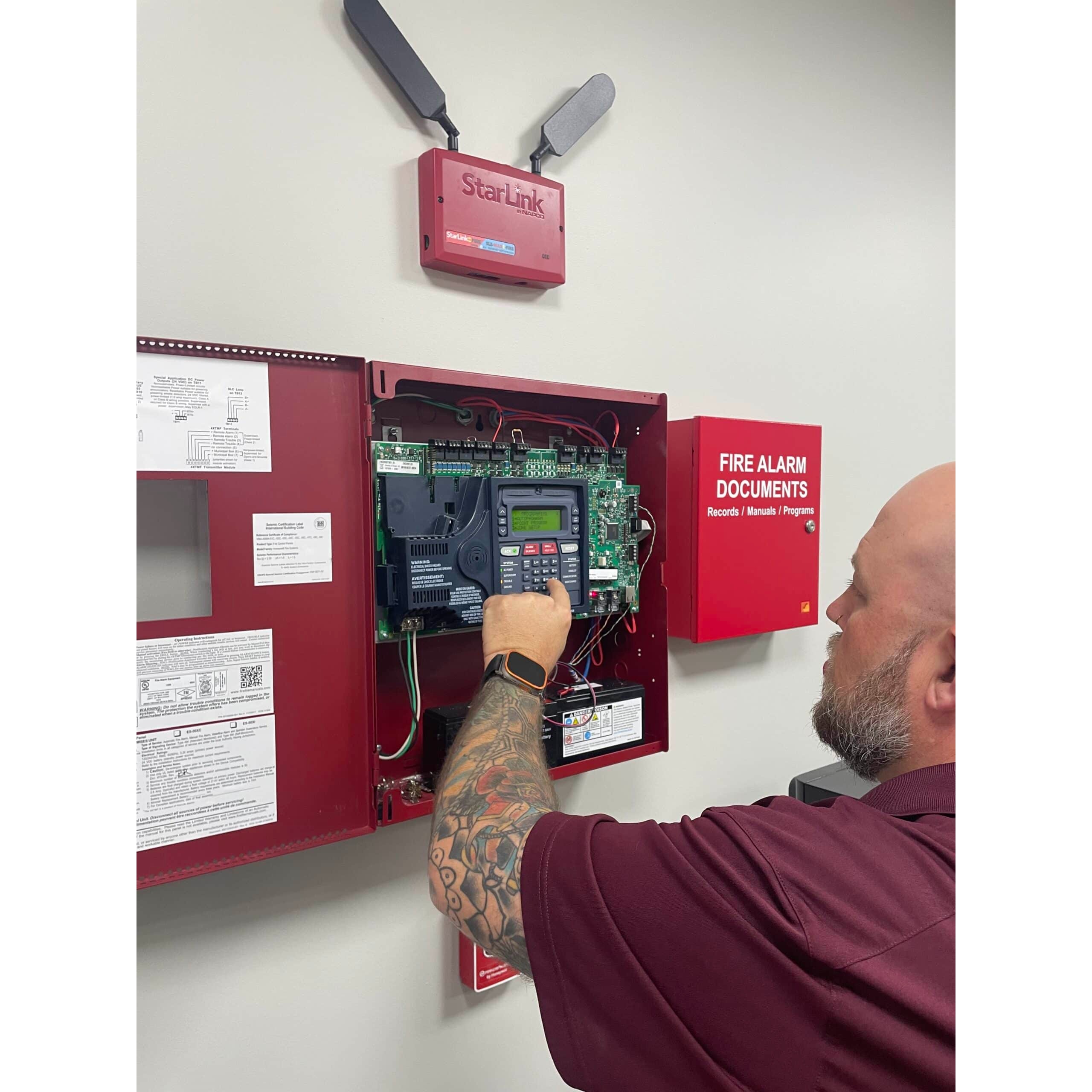 Man with tattooed arm, wearing a maroon shirt, operates a fire alarm panel mounted on a wall, next to a red “Fire Alarm Documents” box.