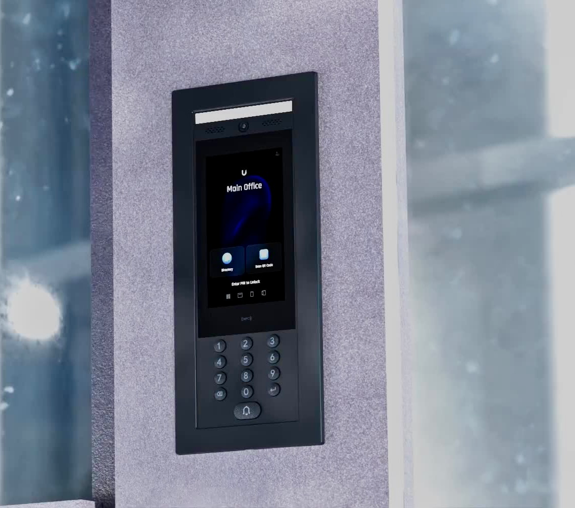 A close-up shot of a modern, black intercom system mounted on a textured gray wall. The intercom features a digital display showing "U Main Office" and options to enter a PIN to unlock. Below the screen is a keypad with numerical buttons and an alarm icon. The background suggests an exterior or a building entrance with a blurred window or glass panel.