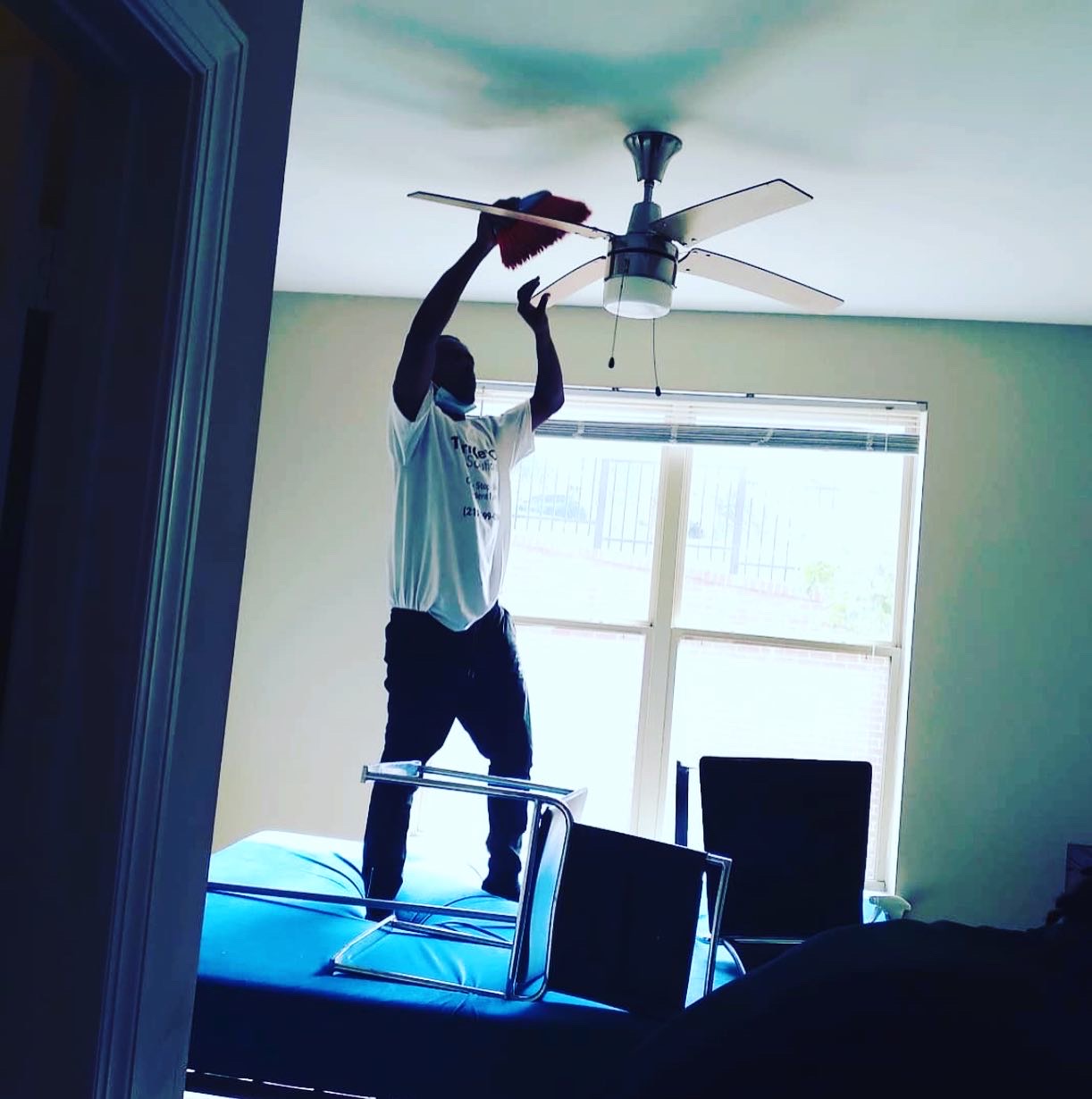 A person wearing a white t-shirt and dark pants is standing on a mattress and cleaning a ceiling fan with a red duster. The person is wearing a face mask. The room has a large window with blinds and two dark chairs are visible. One chair is on its side.