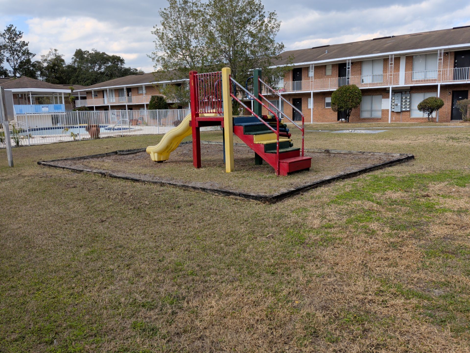 Apartment complex playground with colorful slide and climbing stairs placed on grassy courtyard. Two-story brick residential buildings and fenced swimming pool surround shared outdoor recreation area.
