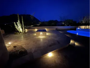 Scottsdale pool and patio at night, Troon North. Travertine decking, built-in gas fire pit, and LED step lighting with the McDowell Mountains in the background. Same project as the pool shot, different angle. Built by Hacienda Outdoors, ROC #325952.