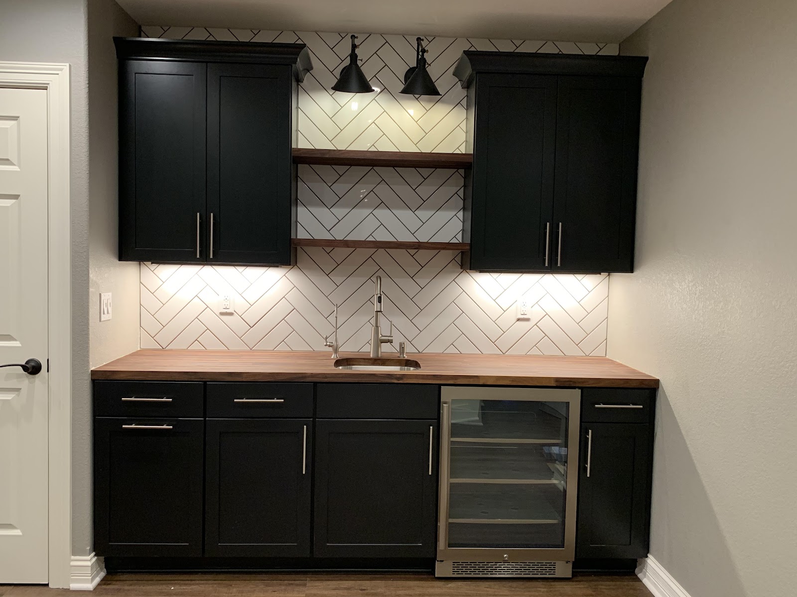 A built-in bar area with black cabinets, a wooden countertop, a stainless steel sink, a wine cooler, and a herringbone tile backsplash. Two black pendant lights are mounted above two floating shelves.