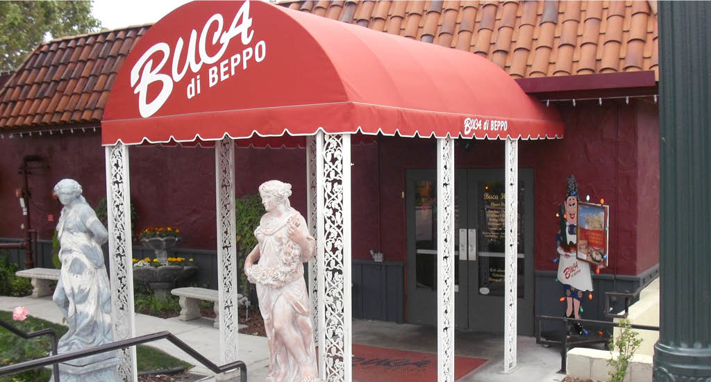 The front entryway with a red cover, featuring Buca signs and statues at Buca di Beppo Claremont.