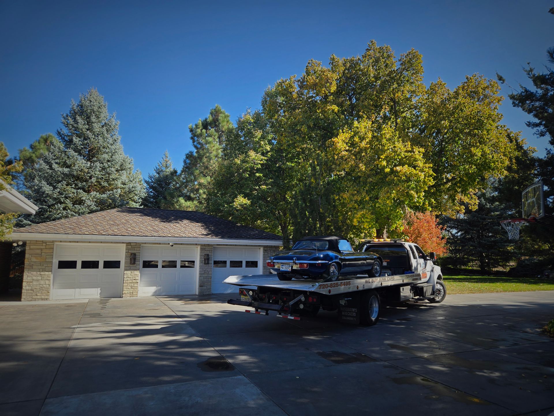 A flatbed tow truck transports a classic car in a residential driveway, surrounded by trees and a garage, showcasing careful vehicle handling and reliable towing services in a calm neighborhood setting.