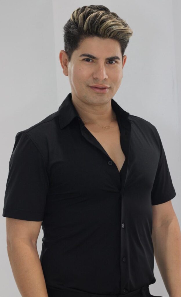 A medium shot portrait of a man with styled blond-streaked dark hair and a black collared shirt. He is looking directly at the camera with a slight smile. He is wearing a delicate gold necklace.