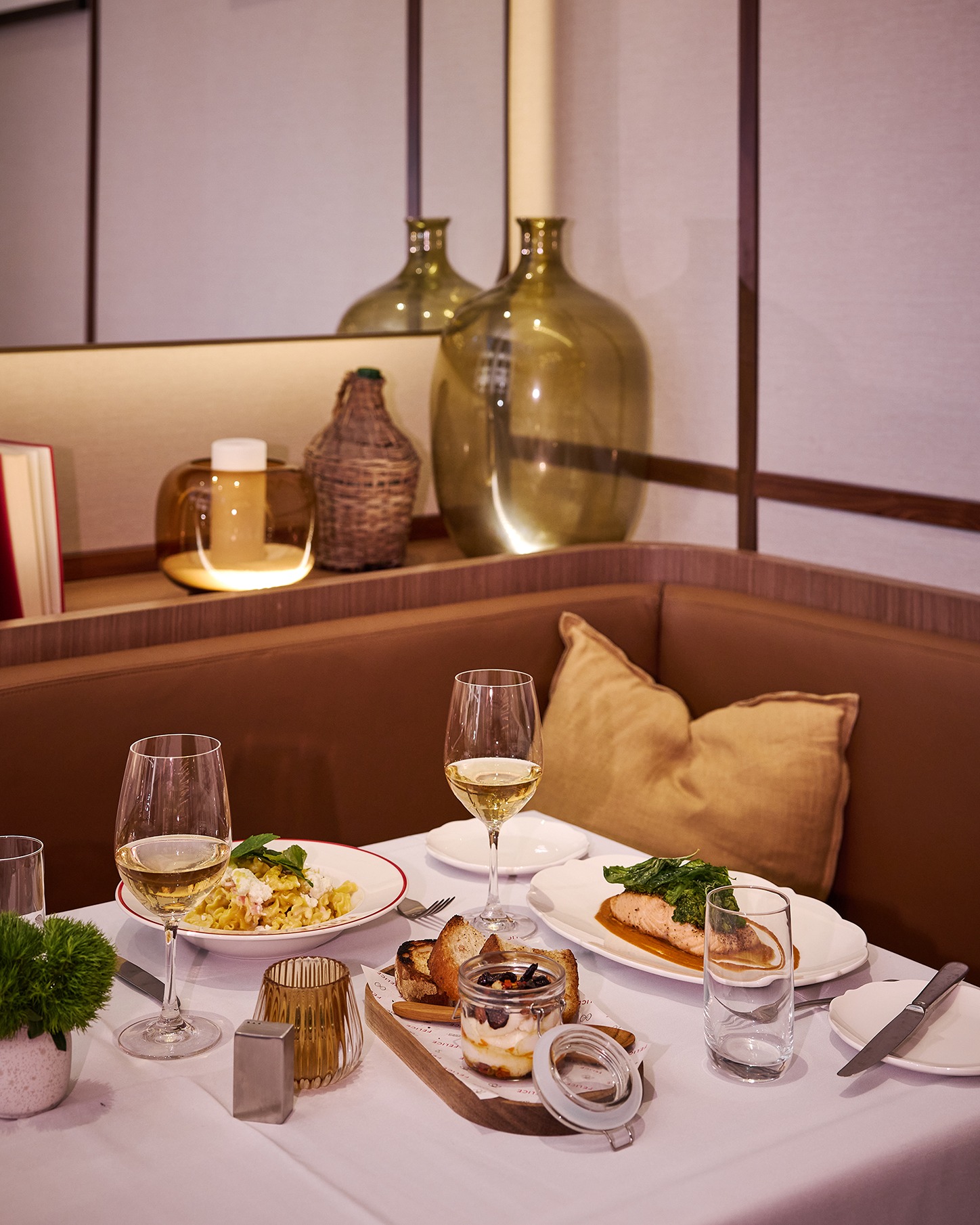A romantic dinner setup featuring white tablecloths, glasses of white wine, a plate of fusilli pasta, grilled salmon, and a tiramisu jar in a warm, wood-paneled booth.