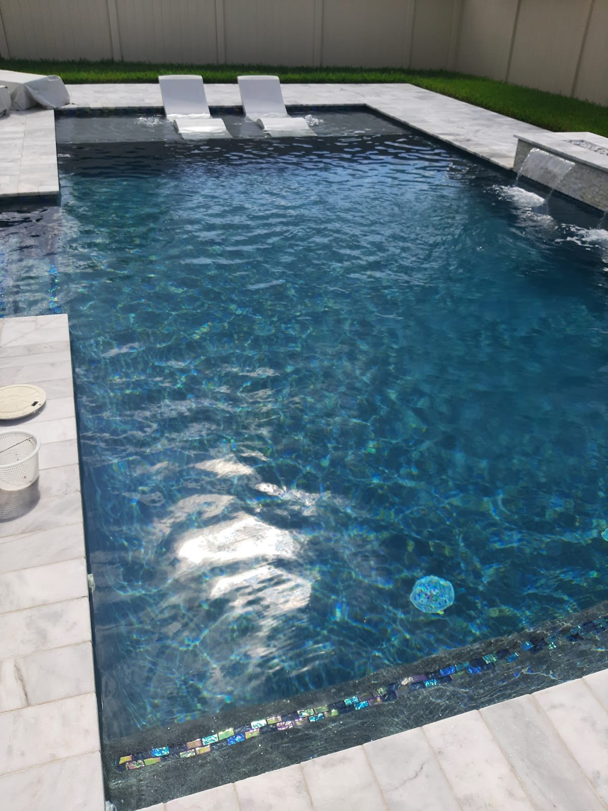 Residential swimming pool with deep blue water, a shallow sun shelf, in-pool lounge chairs, and light stone coping.