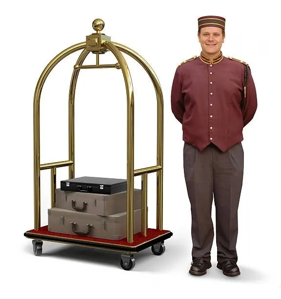 A smiling bellhop stands next to a golden luggage cart loaded with three suitcases. The bellhop is wearing a maroon uniform with gold trim and a matching cap. The luggage cart is made of polished gold metal and has a red carpeted base. The suitcases are stacked on top of each other and appear to be vintage.