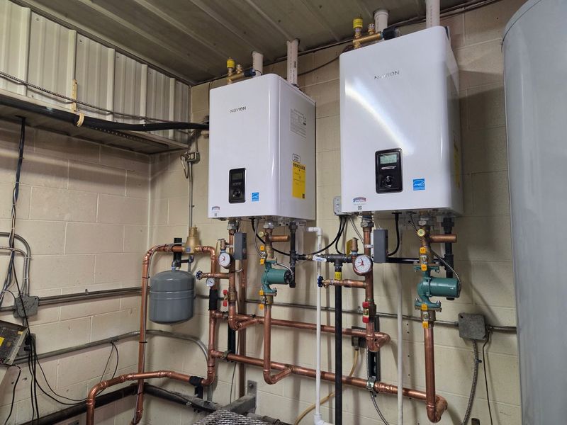 Two white Navien tankless water heaters are mounted on a basement wall, featuring professional copper piping, pressure gauges, and an expansion tank for a high-efficiency residential or commercial heating system.