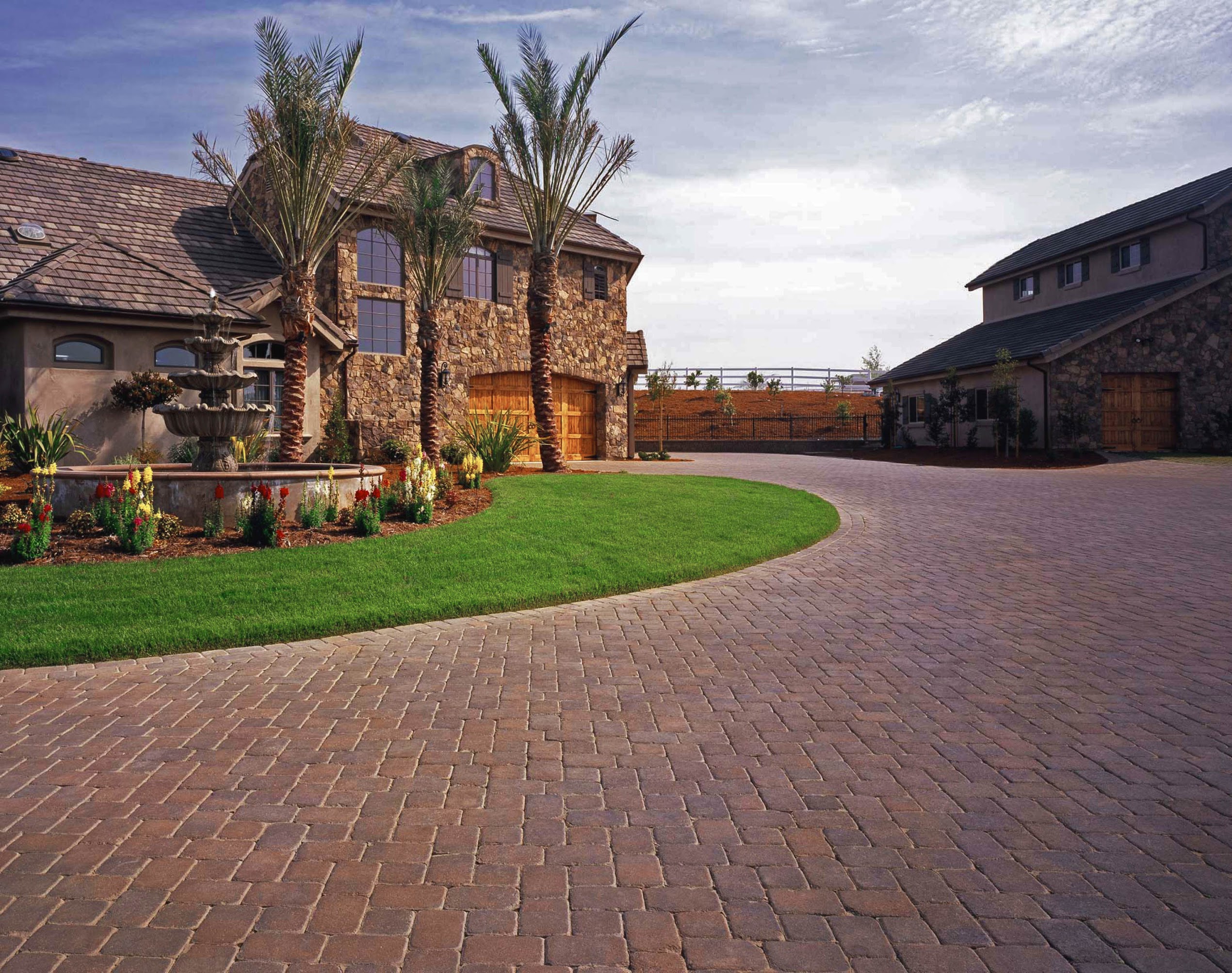 Spacious brick-paved courtyard between rustic stone homes, framed by palm trees, green lawn, flower beds, and a calm blue sky.