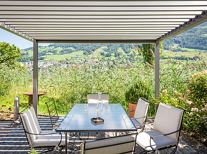 halb-transparente, weiße Standmarkise über einer Terrasse mit Glas-Tisch und Stühlen, inmitten der Natur.