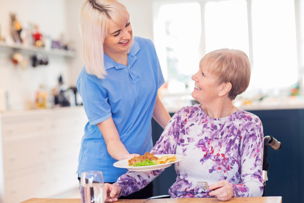 A young woman in a blue polo shirt is serving a plate of food to an elderly woman sitting in a wheelchair. The elderly woman is wearing a purple and white floral top and is smiling. The scene appears to be a home kitchen or dining area, with bright natural light coming from large windows in the background. The food on the plate includes what looks like meat, peas, and some kind of flatbread or pastry.
