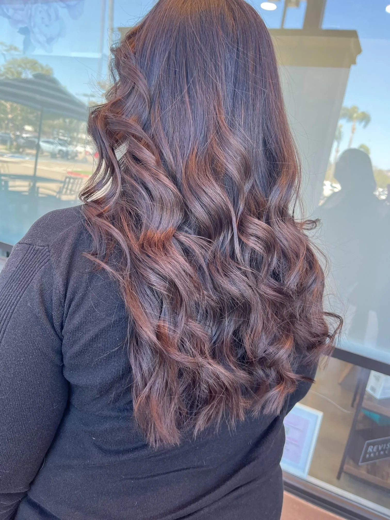 Long dark brown hair styled in soft curls with subtle reddish highlights, creating rich dimension and shine. The hairstyle is photographed from the back to highlight professional salon coloring and styling.