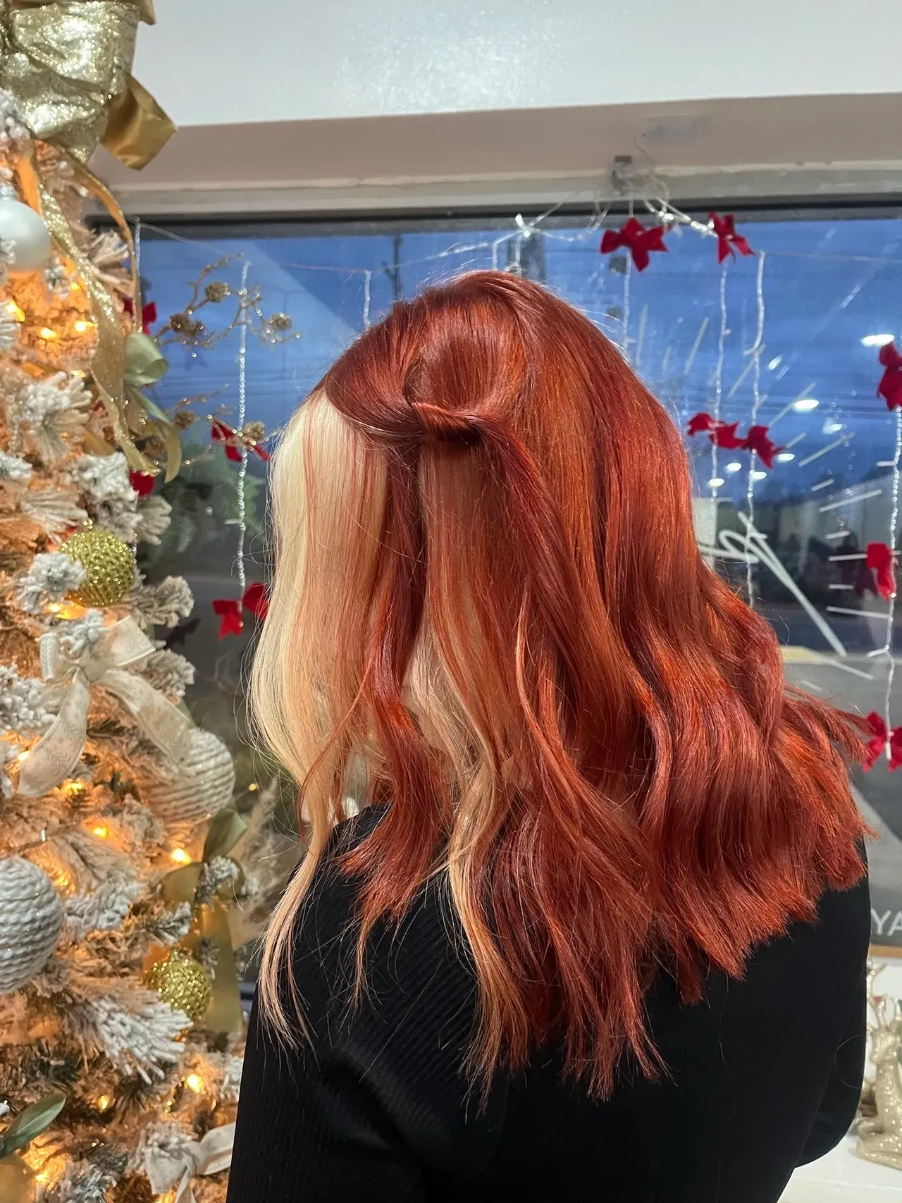 Woman showcasing vibrant copper red hair with blonde front section styled in soft waves, photographed indoors near decorated Christmas tree.