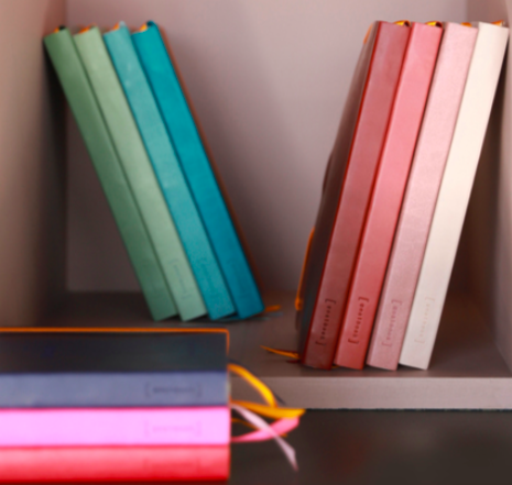 A professional product image by Tabula Rasa Social Stationers, displaying a vibrant array of Rhodia Goalbooks. The photograph features numerous notebooks in new 2021 colors, including Nacarat, Rose Smoke, Celadon, Aqua, Sage, and Peacock, arranged on shelves to highlight their appealing palette and minimalist design. These Rhodia journals epitomize French quality, crafted with ultra-smooth, acid-free, and fountain pen-friendly paper, providing an ideal surface for bullet journaling, planning, and capturing thoughts with precision and refinement.