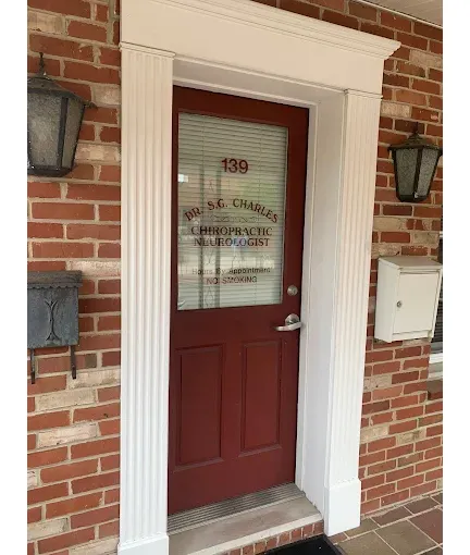 It&rsquo;s a photo of the front entrance to a brick building with a dark red/maroon door framed by white trim and columns. There are two black wall lantern lights on either side of the doorway, plus a mailbox mounted on the right and a small wall-mounted box/fixture on the left.
On the glass window of the door, there&rsquo;s lettering that includes the street number &ldquo;139&rdquo; and the practice name/details, including &ldquo;Dr. S.G. Charles&rdquo; and &ldquo;Chiropractic Neurologist,&rdquo; along with a note about appointments (and a line indicating no soliciting).