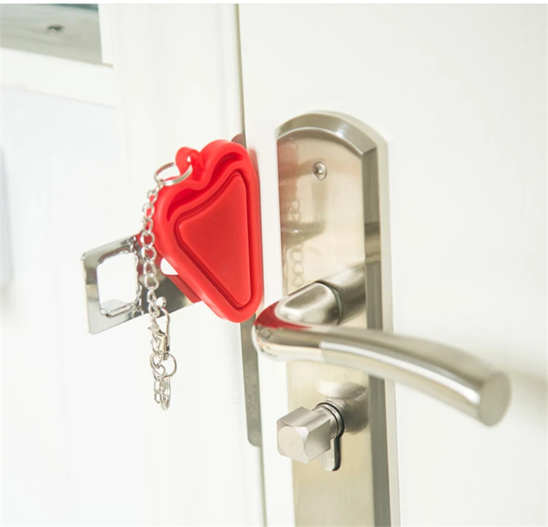 A red triangular door stop is attached to a silver door handle and lock mechanism. The door stop is made of plastic and has a chain attached to it. The door handle is a lever-style handle made of brushed metal. The lock mechanism is a deadbolt with a hexagonal keyhole. The door is white.