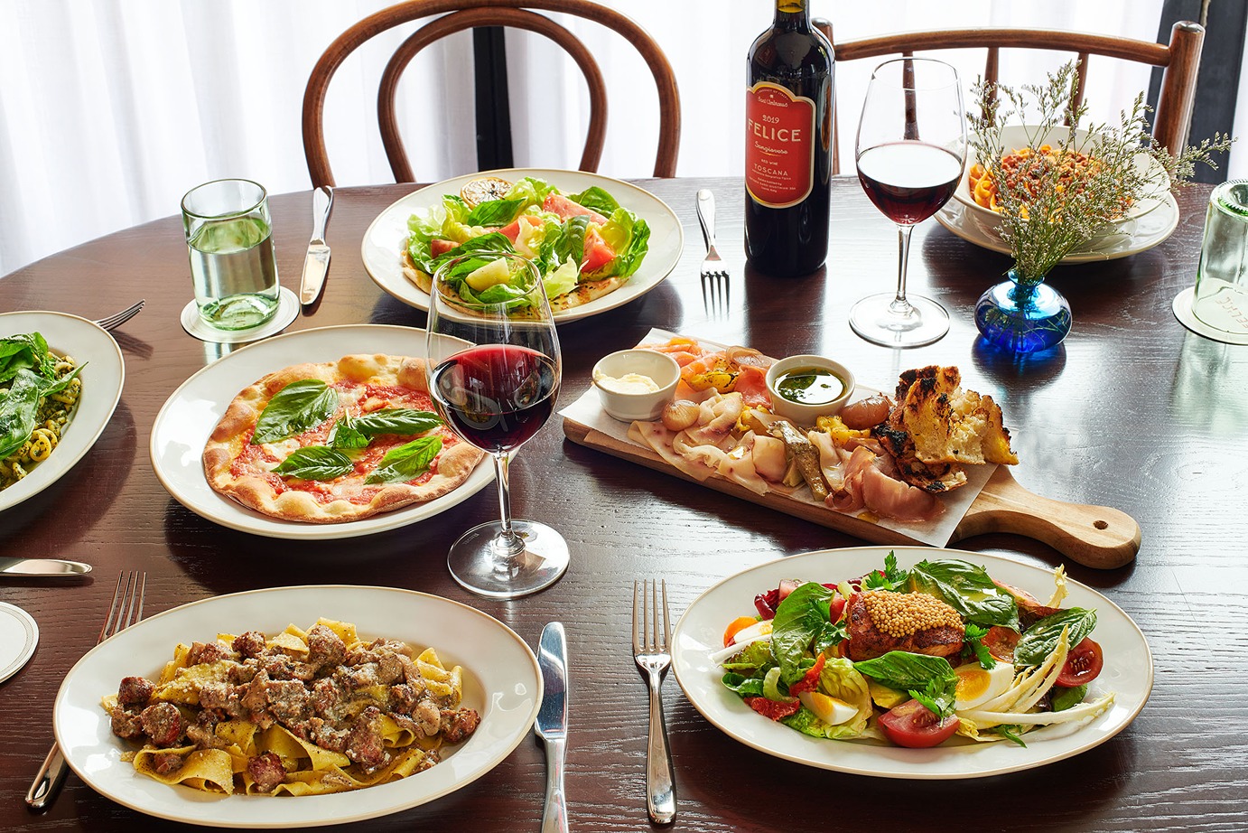 A festive spread featuring a margherita pizza, pasta with meat sauce, fresh salads, and a wooden board of Italian meats and cheeses, accompanied by red wine.