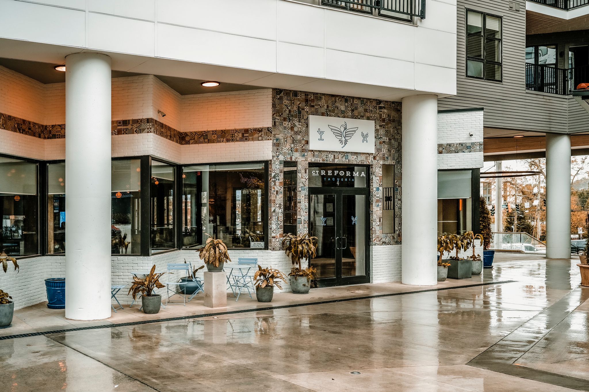 The building exterior highlights symmetry and detail, with white brick walls and patterned tiles. Large windows offer a welcoming glimpse of the interior. The outdoor space is clean and polished, with modern planters and casual seating adding charm