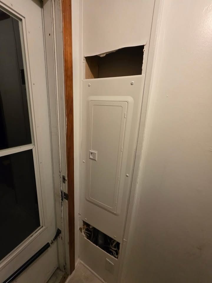 Electrical panel door installed in wall with open sections exposing wiring behind, located near doorway, showing incomplete finishing or access openings for electrical components in residential interior space.