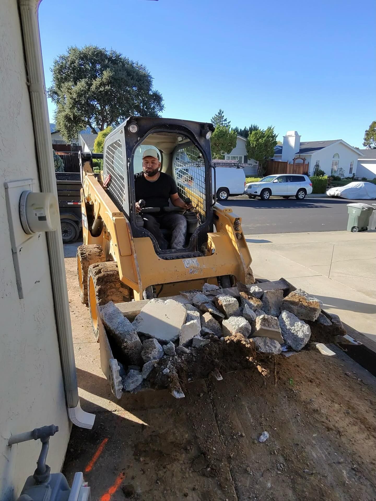 Worker operating a skid steer loader removing broken concrete and debris from a residential driveway, showcasing excavation work, heavy equipment use, and site preparation in a suburban neighborhood under clear daylight.