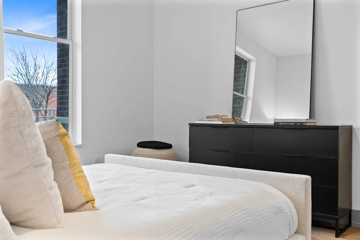 A bedroom with a white bed, a black dresser, and a large mirror. 