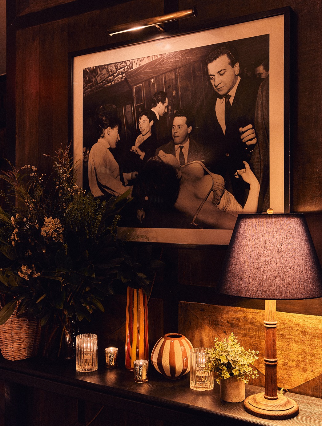 A moody corner featuring a large framed photo of people at a party illuminated by a picture light. Below a table holds a blue lampshade flickering candles and a striped vase.