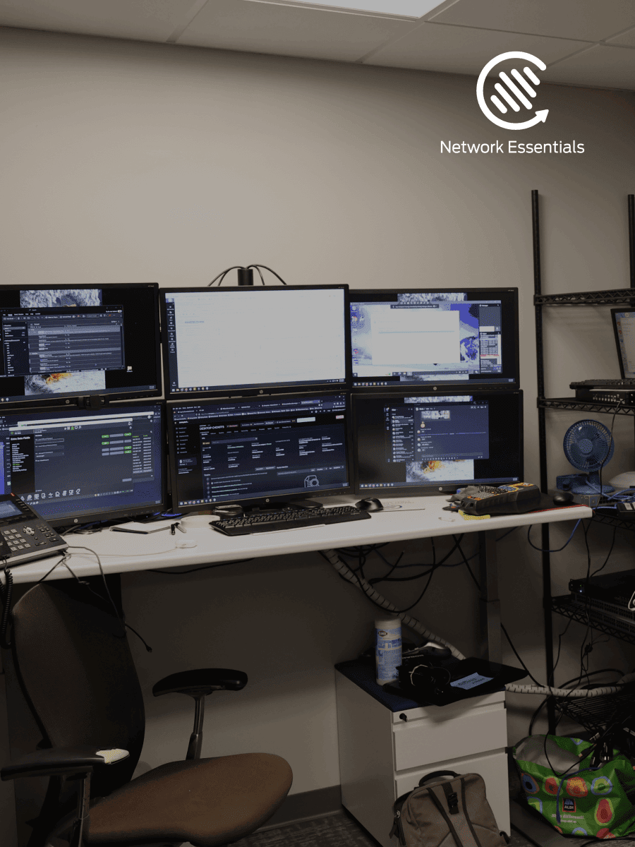 A dark room with multiple computer monitors set up on a desk, displaying complex data. A chair is positioned in front of the desk, and a rolling cabinet is placed to the side. The room also features shelving units stacked with equipment, a fan, and a bag. The logo of 'Network Essentials' is visible in the upper right corner.