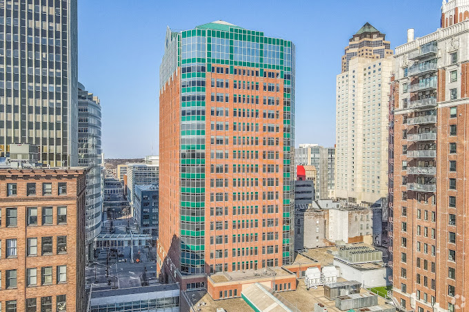 A vibrant cityscape featuring a tall, distinctive red-brick building with green-tinted windows and a modern geometric design. The building is surrounded by other high-rise structures of varying architectural styles, including classic brick facades and contemporary glass towers. The bright blue sky enhances the urban skyline, reflecting a clear, sunny day in a bustling downtown area.