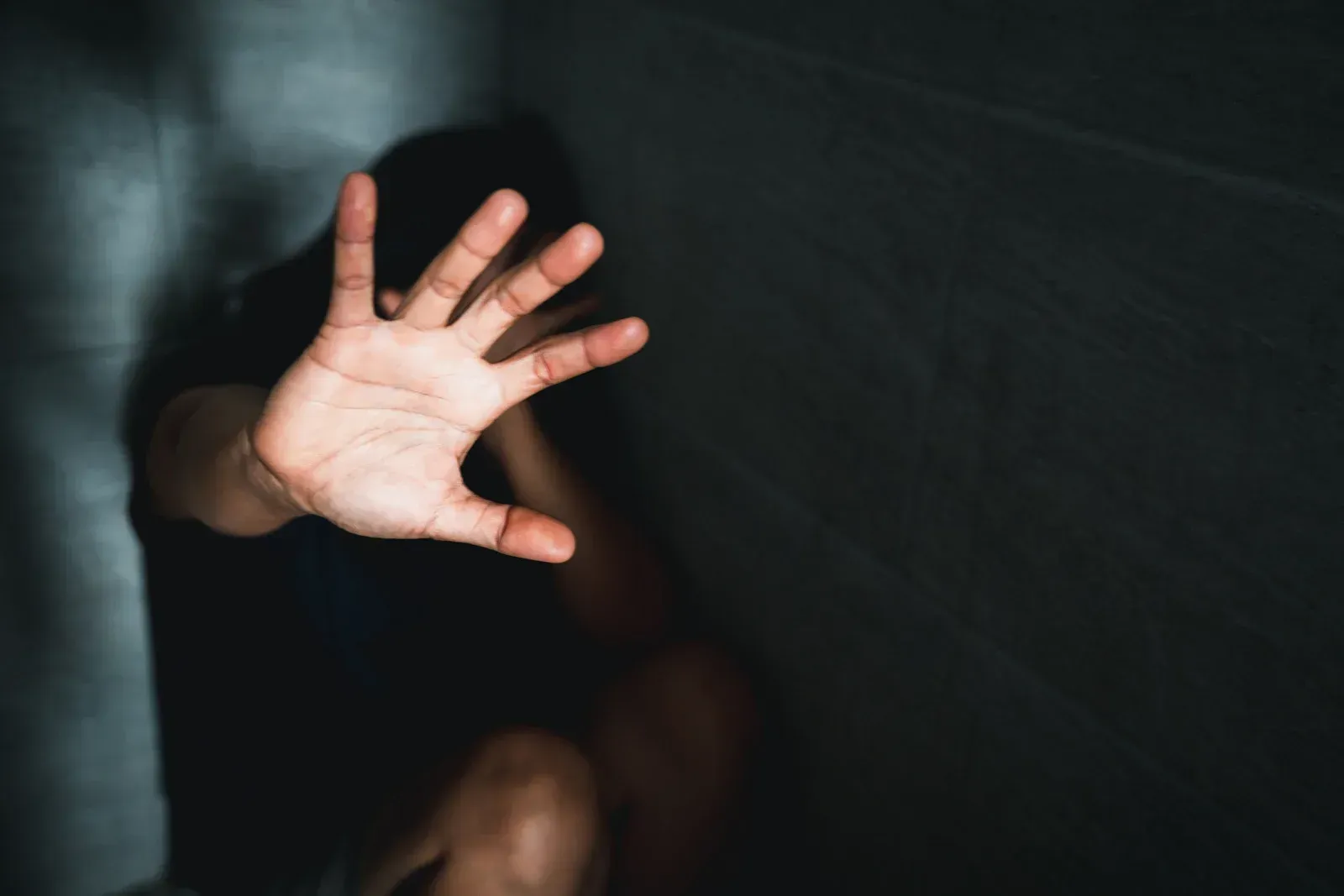 A dimly lit image shows a person with their hand outstretched in a gesture of stopping or defense. The person's face is obscured by shadow and their hand. The background is dark and textured, possibly a wall.