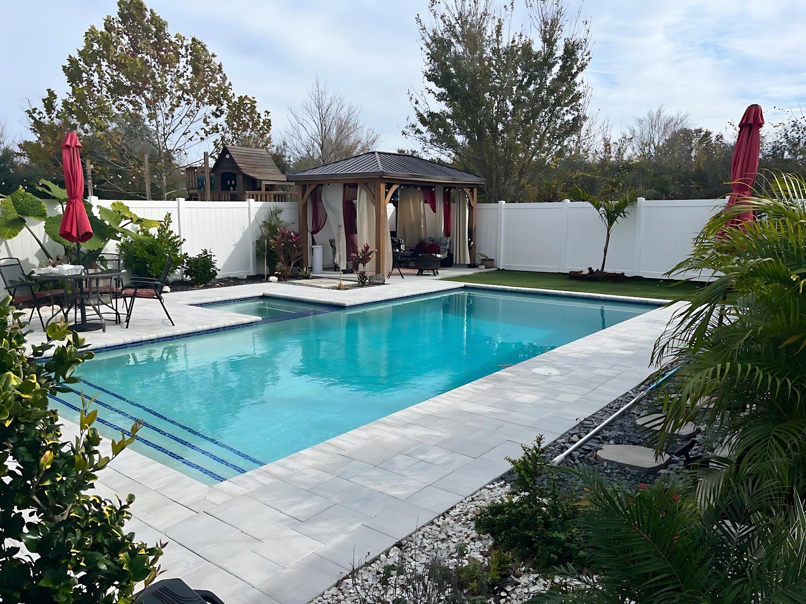 Rectangular residential swimming pool with clear blue water, light stone decking, tropical landscaping, and an outdoor cabana area.