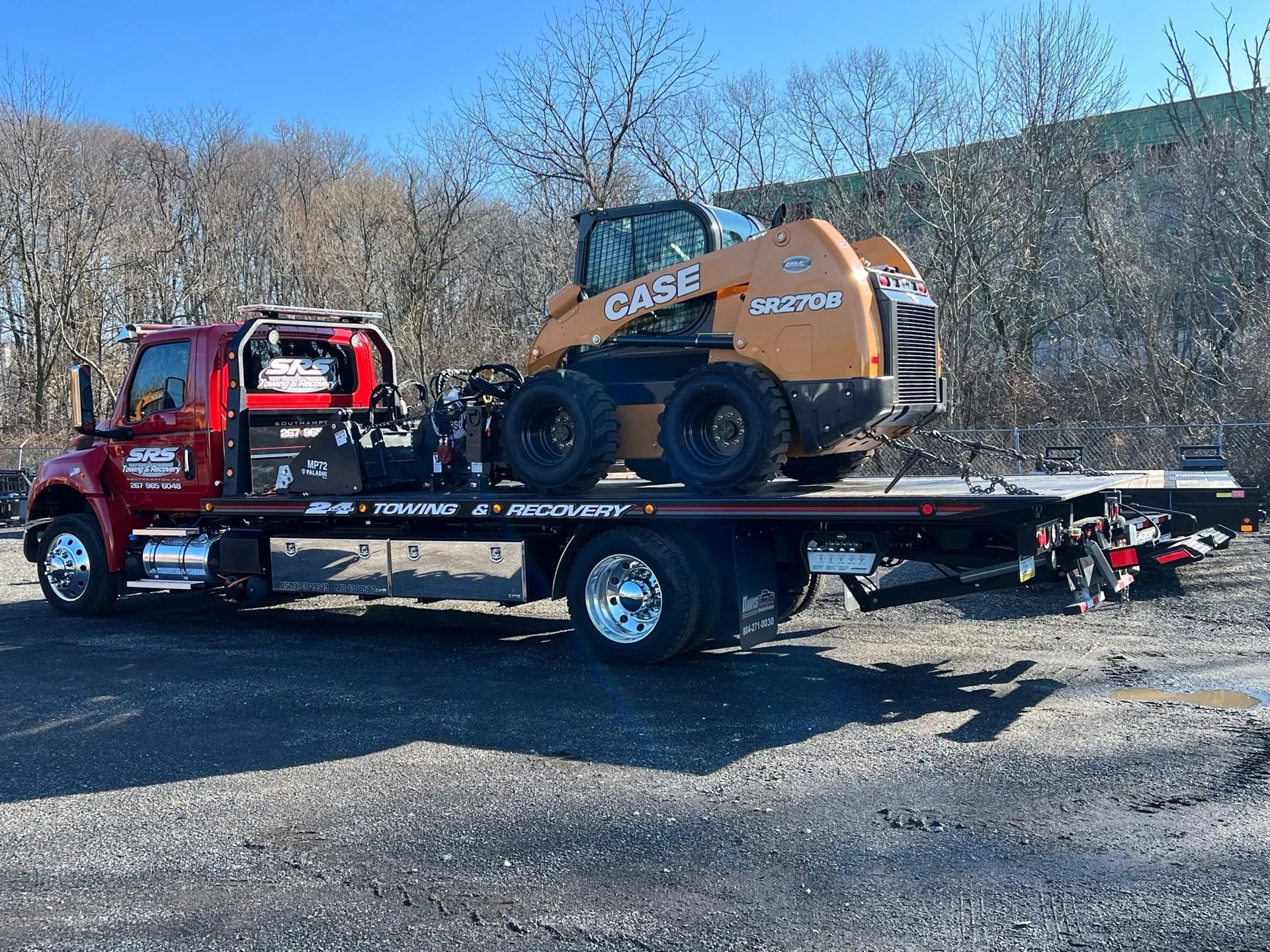 A red SRS Towing & Recovery flatbed truck transporting a CASE SR270B skid steer loader in daylight. The machine is secured with chains, and the truck's polished chrome details reflect sunlight. Leafless trees and an industrial building are visible in the background.