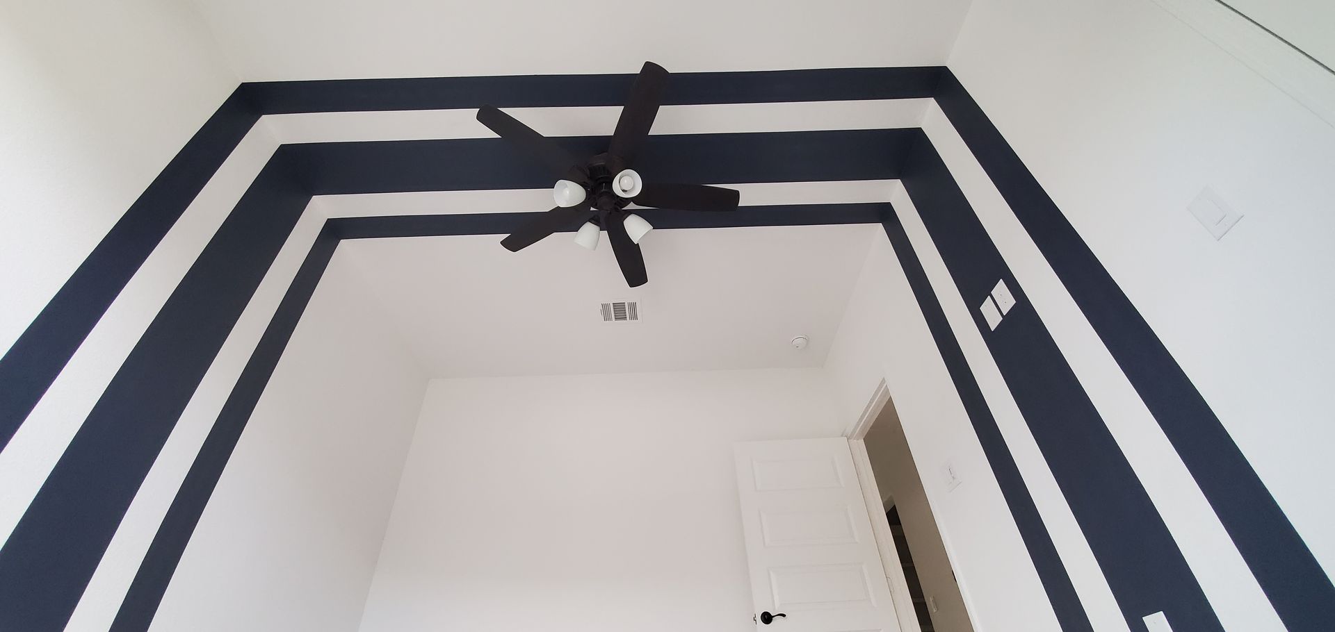 Spacious bedroom showcasing bold black painted geometric ceiling accents framing the room, complemented by white walls, ceiling fan, modern trim, and clean architectural lines for dramatic visual appeal.
