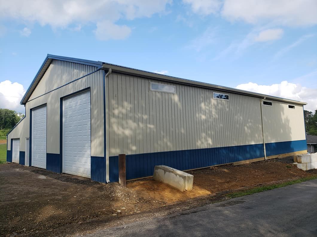 A large metal garage building with beige siding and blue lower panels, featuring tall white roll-up doors, small horizontal windows, and installed gutter system, positioned on a sloped gravel and dirt area.