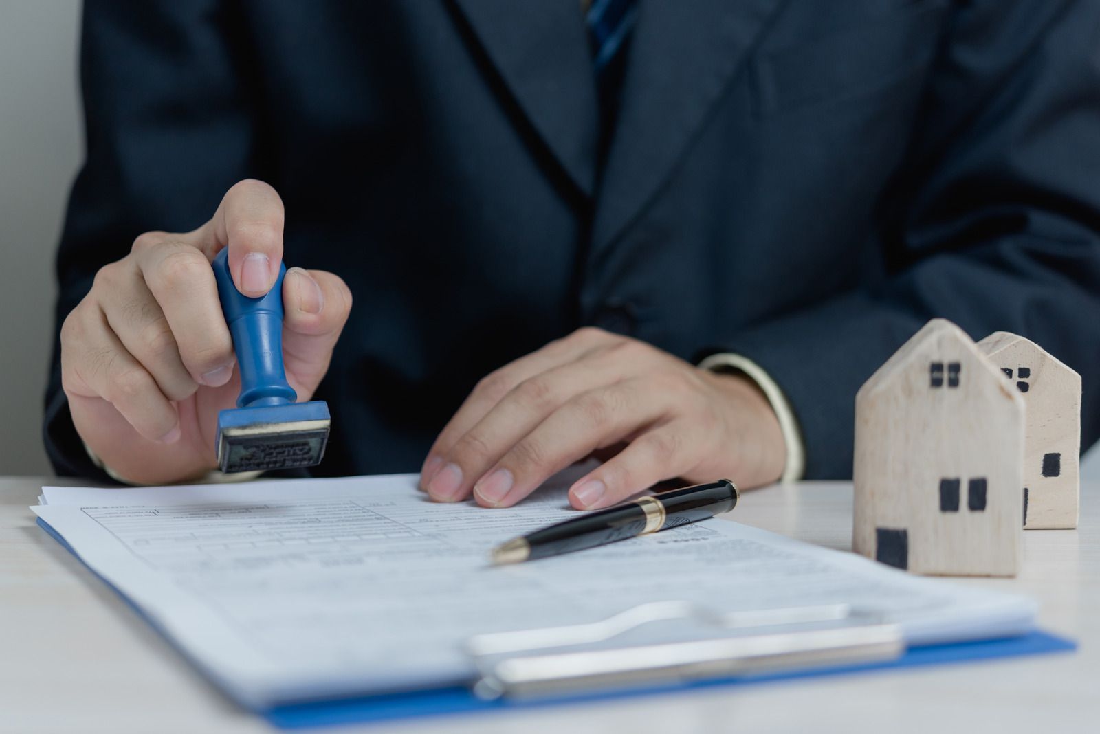 Business professional stamping documents with miniature house model nearby, representing property registration, legal verification, real estate documentation, and official approval process required for secure property transactions and ownership confirmation.