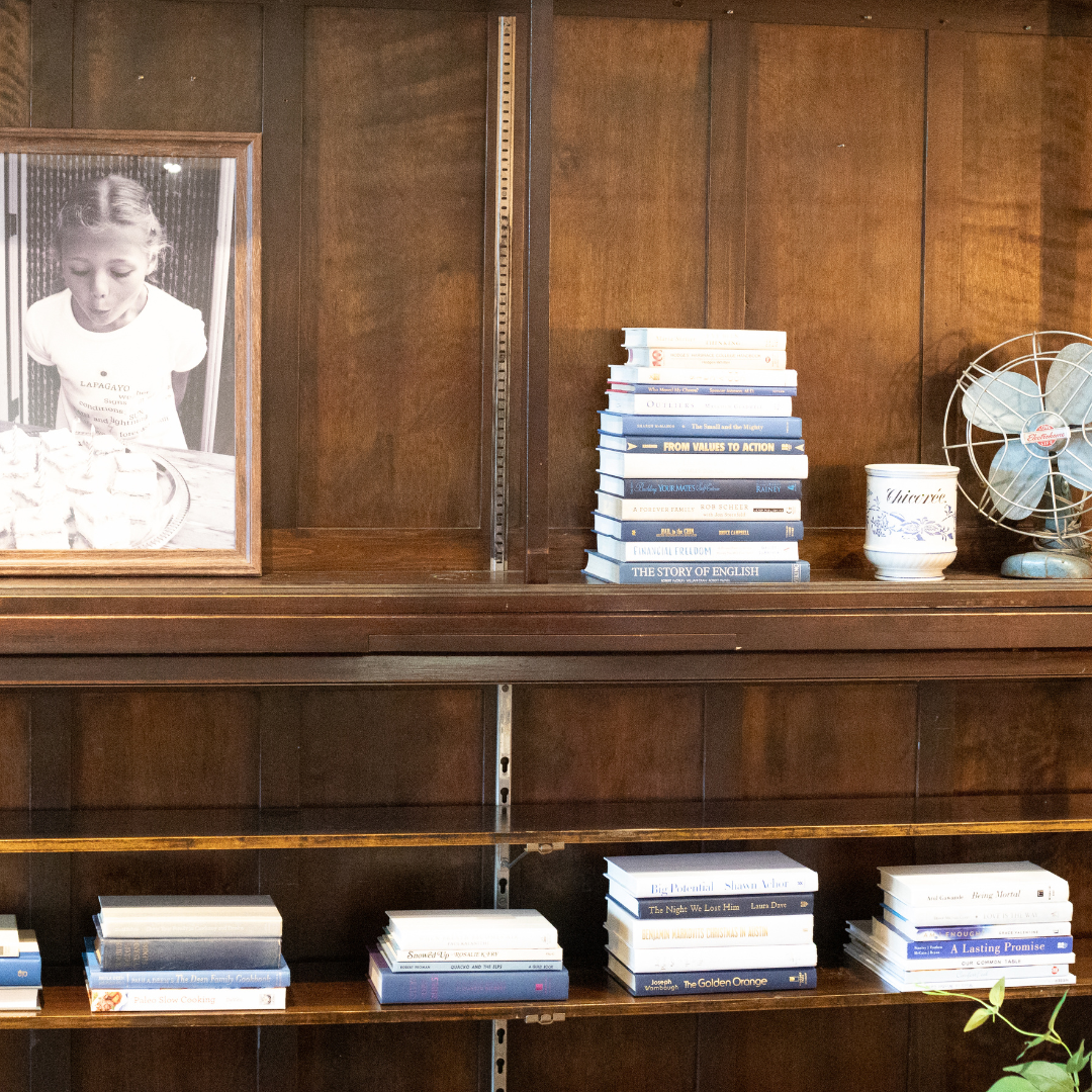 A wooden bookshelf against a wood panel backdrop. There is a framed black and white photograph of a  