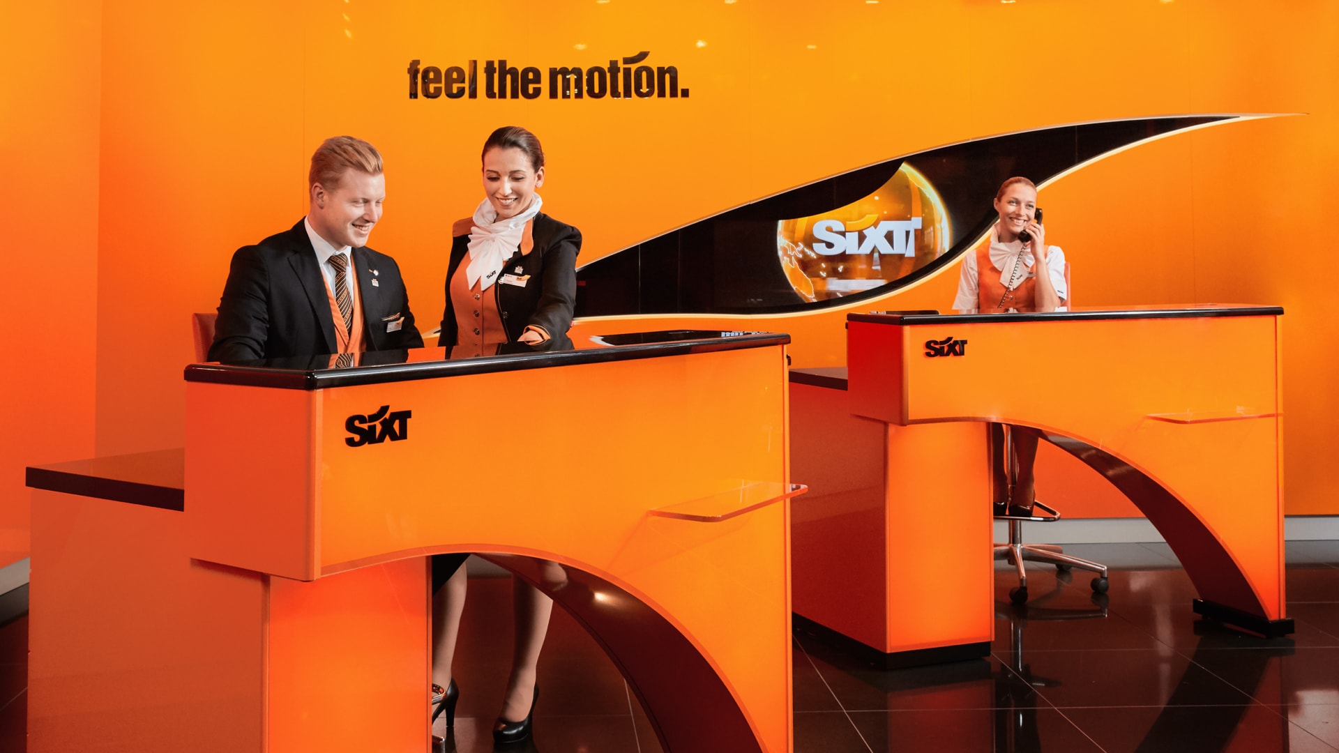 Three Sixt employees dressed in black and orange business attire work in front of an orange wall with "feel the motion" written on the wall in black.