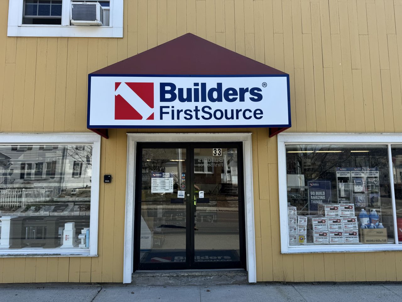 Front entrance of a Builders FirstSource location with a branded awning sign, glass double doors, and window displays on a yellow building materials storefront.