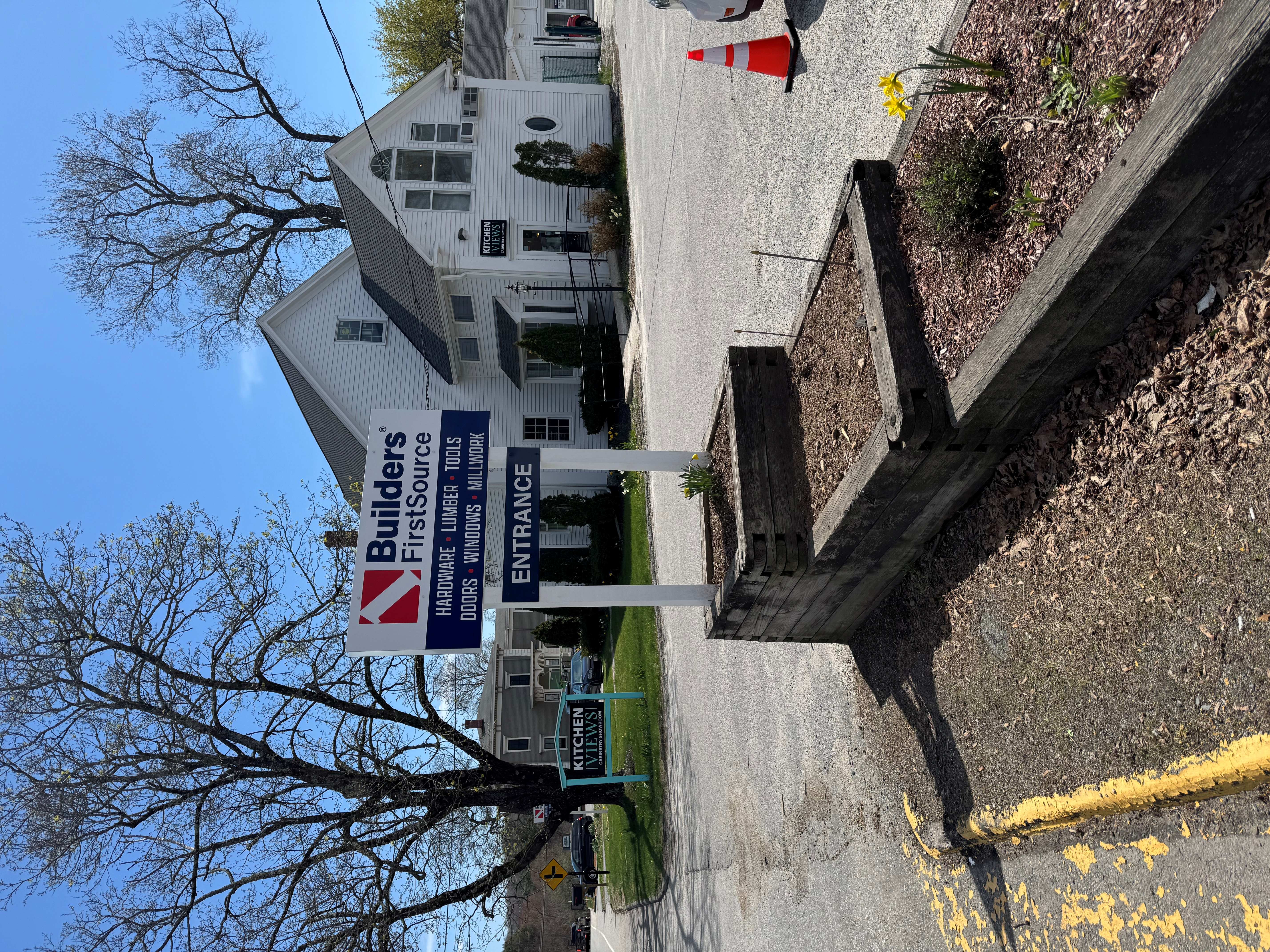 Builders FirstSource roadside sign listing hardware, lumber, tools, doors, windows, and millwork, positioned at the gravel entrance to a white building with trees and planters in front.