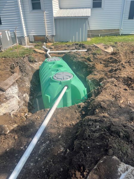 A high-angle view of a new, bright green plastic septic tank installed in a deep dirt trench behind a white house. White PVC pipes are connected to the system.