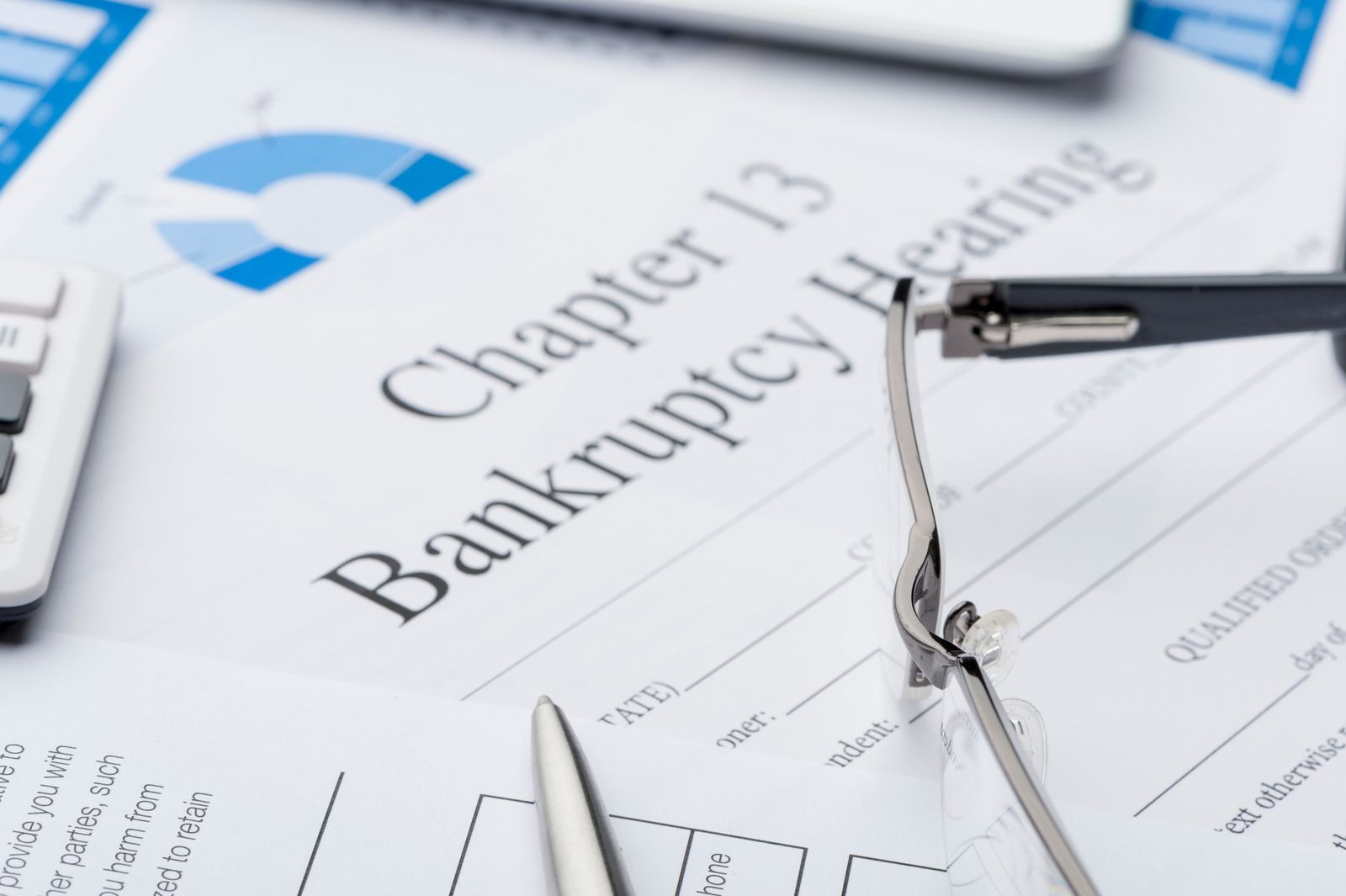 A close-up of a Chapter 13 Bankruptcy Hearing document on a desk, with eyeglasses and a silver pen resting on top, and a calculator and blue financial charts nearby.