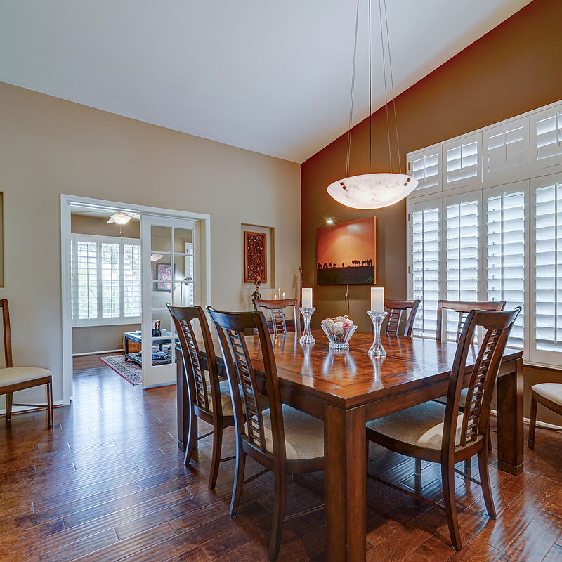 A formal dining room is set with a large wooden table and chairs, illuminated by a modern pendant light. The room features warm brown walls, white plantation shutters on the windows, and hardwood floors. A painting of a sunset with silhouetted figures hangs on the wall, and decorative candles and a bowl of fruit adorn the table.