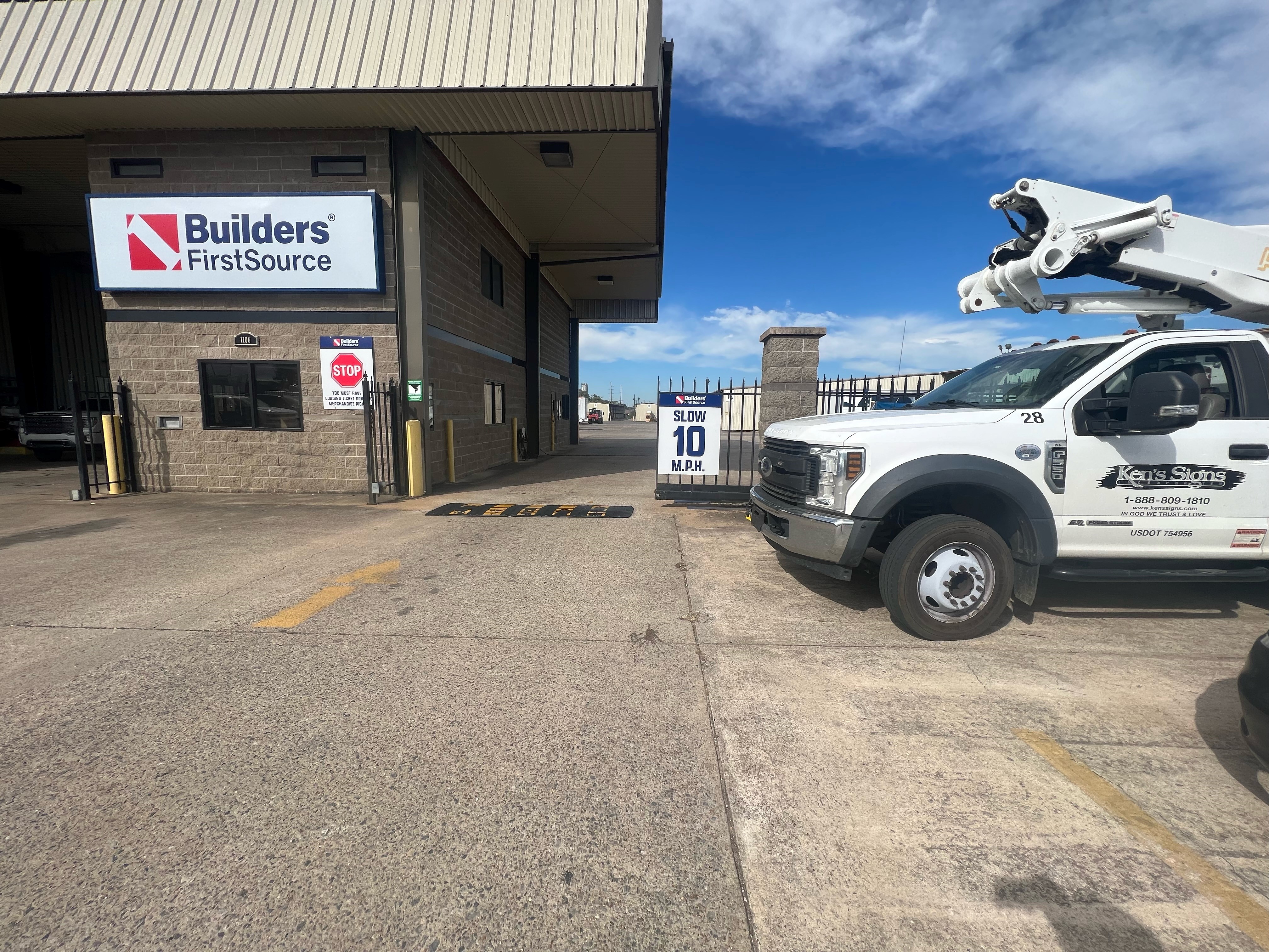 A receiving area at a Builders FirstSource facility with a large company sign mounted above the entrance. A "STOP" sign and instructions are visible near a window. To the right, a white work truck with "Ken's Signs" branding is parked. A sign indicating "Slow 10 M.P.H." is posted near the entrance, and the area appears to be designed for trucks entering and exiting the yard. The sky is partly cloudy with blue skies visible above.