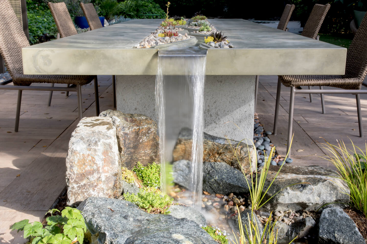Outdoor dining table with a built-in waterfall spilling into a rock garden, surrounded by woven chairs, succulents, and greenery.