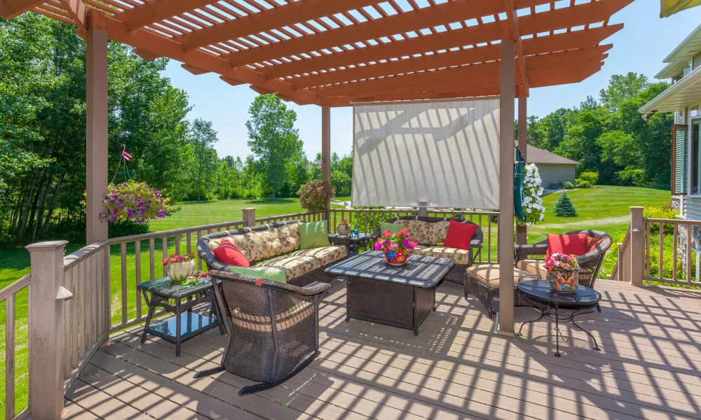 A beautifully decorated outdoor deck with a wooden pergola, comfortable wicker furniture, and vibrant floral arrangements. The deck overlooks a lush green lawn and mature trees under a clear blue sky, suggesting a peaceful and inviting outdoor living space.