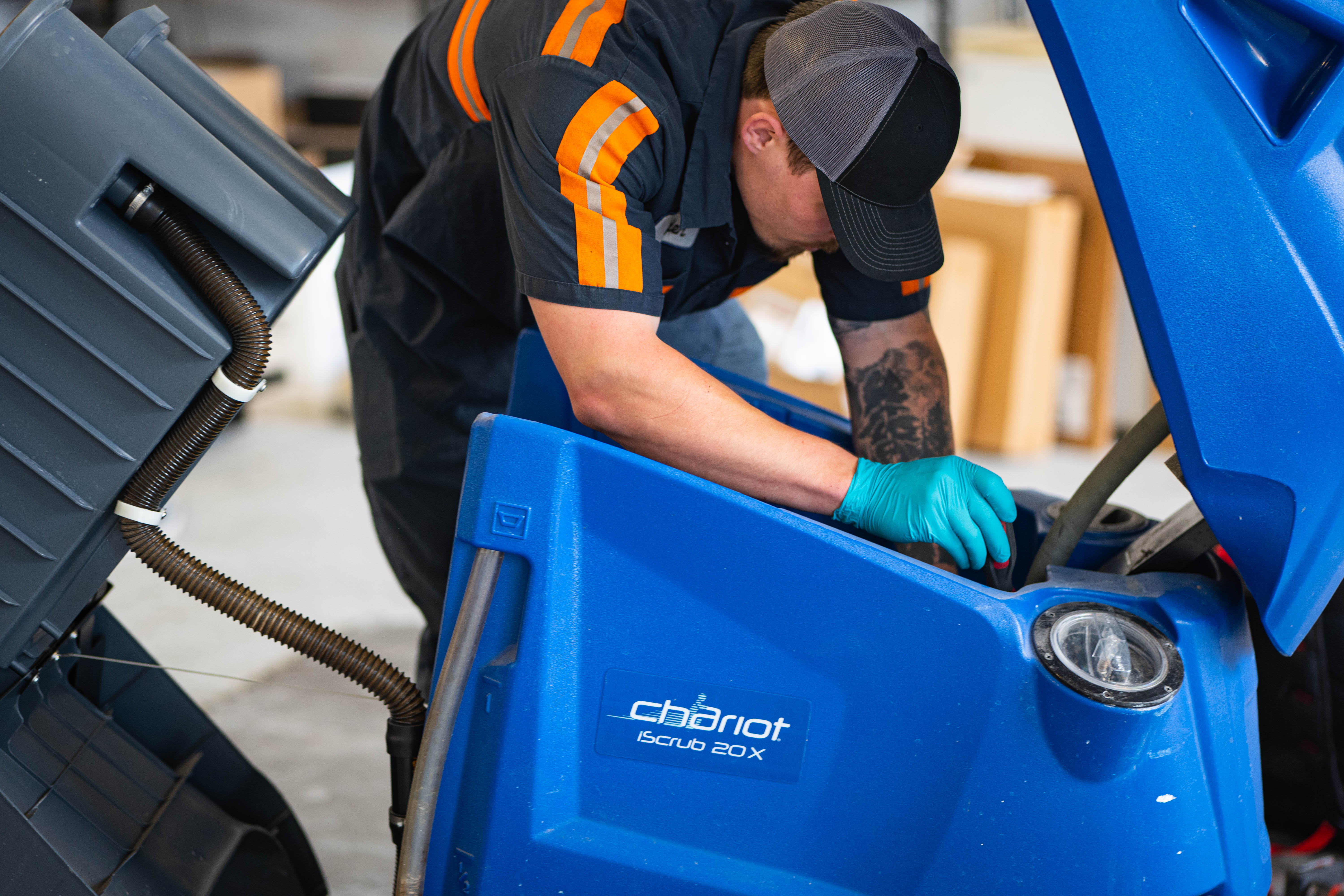 A WaySolutions technician in a high-visibility service uniform reaches into the open internal compartment of a blue Kärcher Windsor Chariot iScrub 20X. The shot shows the machine's recovery tank tilted back, allowing for deep-level mechanical inspection and hose maintenance. The technician is wearing teal nitrile gloves for safety and precision.